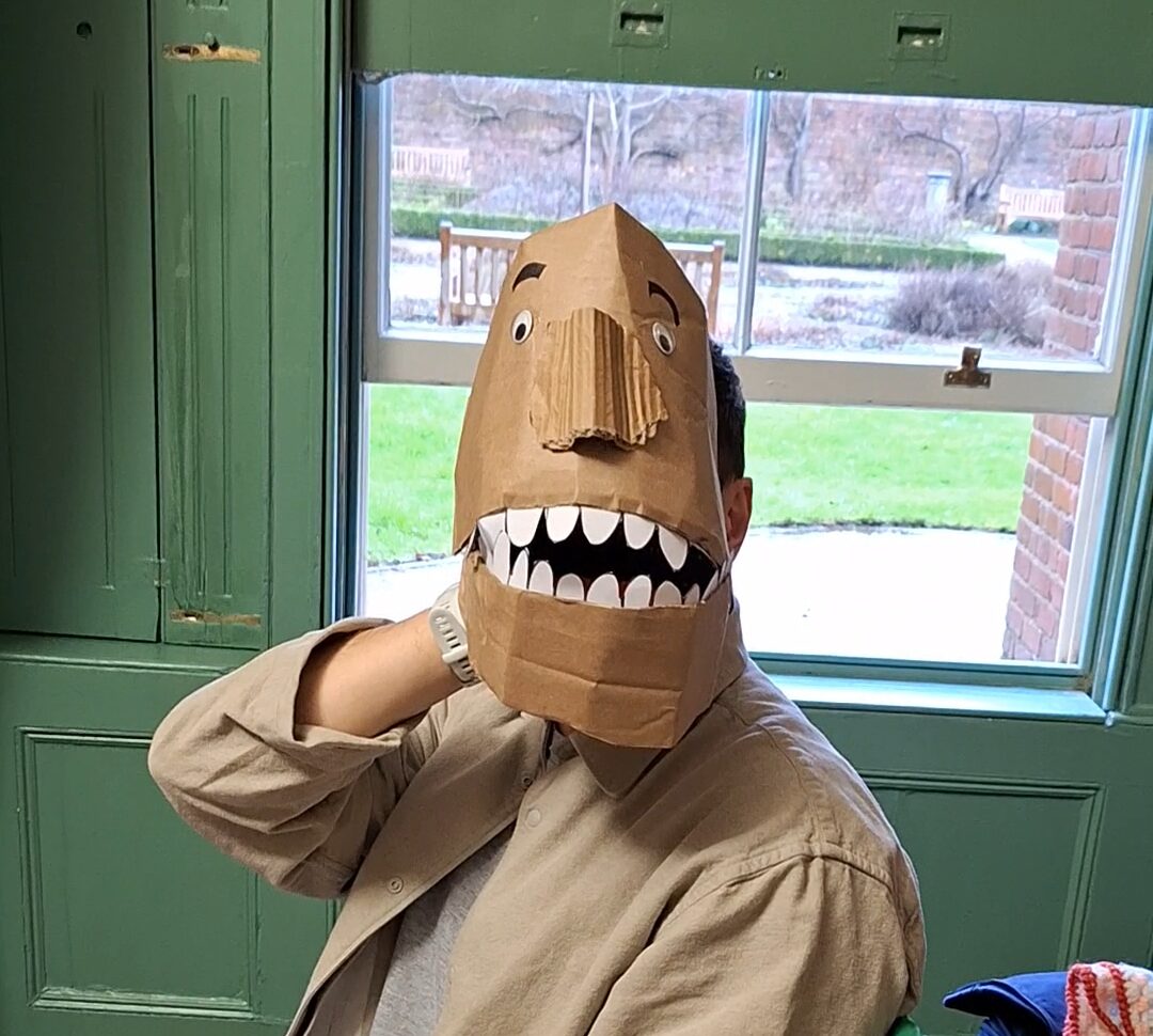 A person wearing a brown paper mask with a wide mouth and teeth drawn on it sits indoors near a window, with green walls and a view of grass and bushes outside.
