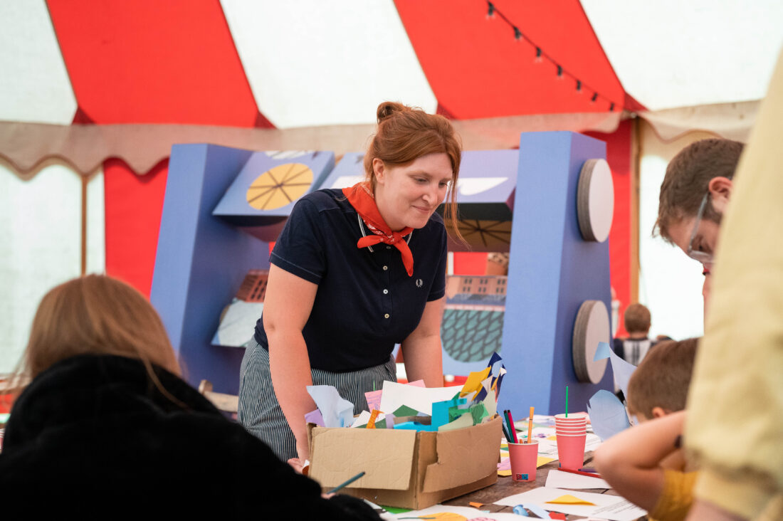 A woman with a red scarf stands by a table covered in colourful paper, helping children with a craft activity inside a marquee with a red and white striped ceiling.