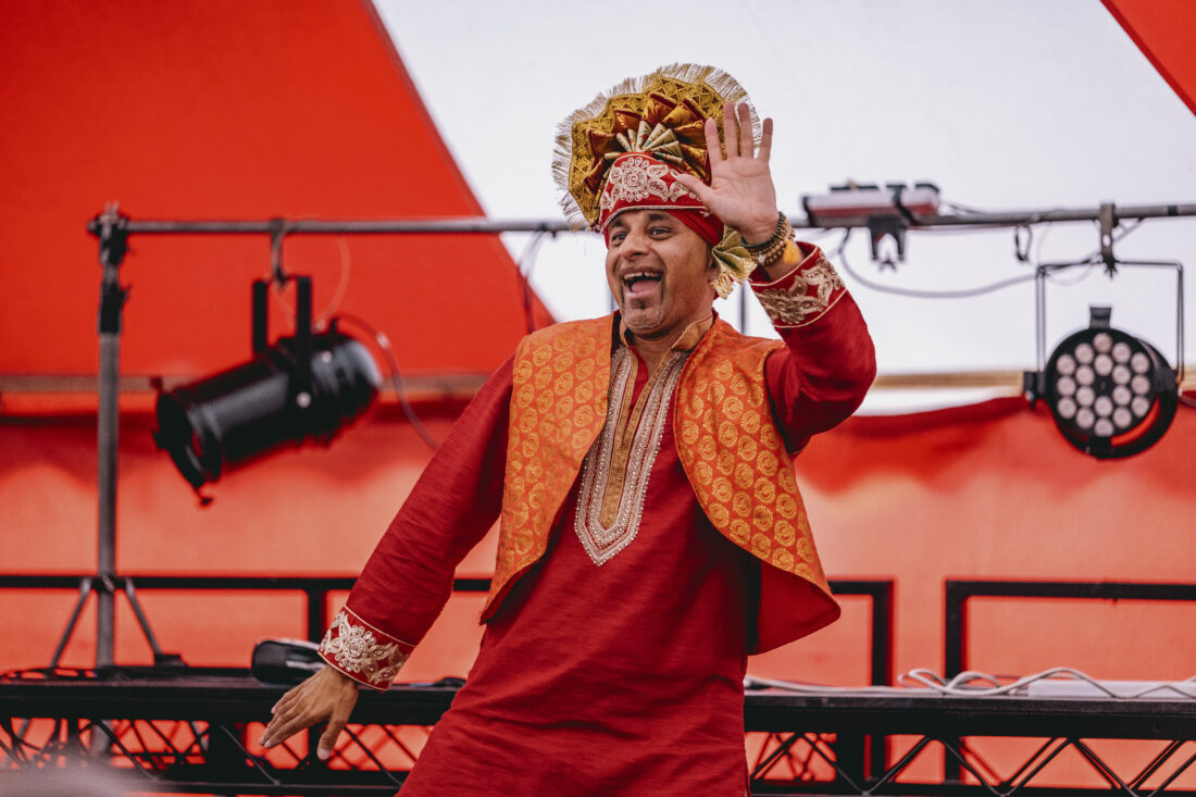A man in a red traditional outfit and ornate headpiece smiles and waves on stage, with red and white striped background and stage lights behind him.