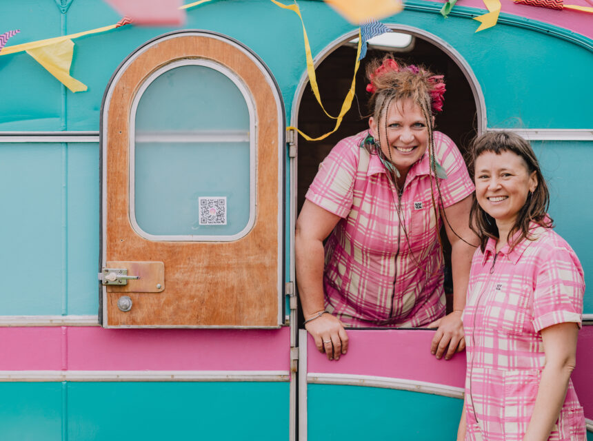 Two smiling women in pink checked dresses stand by a colourful teal and pink caravan decorated with bunting. One woman leans out of the door, while the other stands beside her outside.