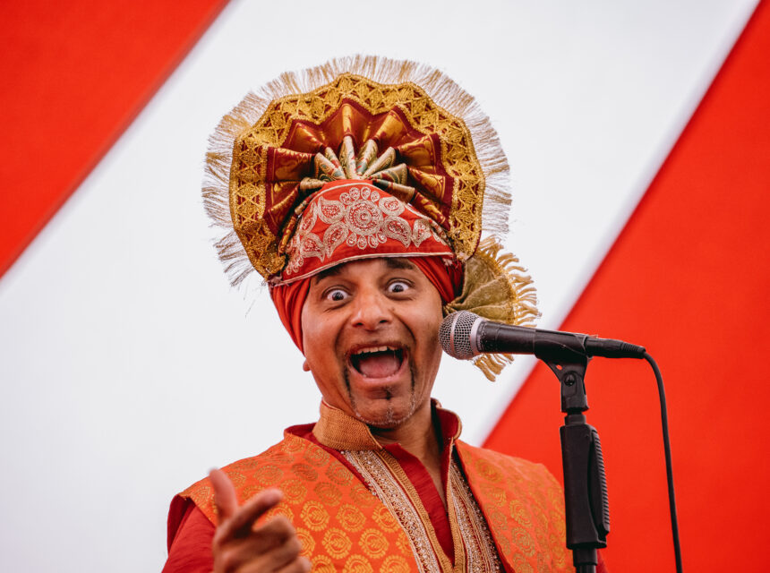 A person wearing an ornate, gold and red traditional headdress and outfit enthusiastically speaks or sings into a microphone, gesturing and smiling with a red and white striped background behind them.