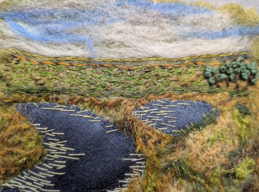 An embroidered landscape shows a blue river winding through grassy fields, with green trees to the right and a cloudy sky above. Various threads create texture for grass, water, and sky.