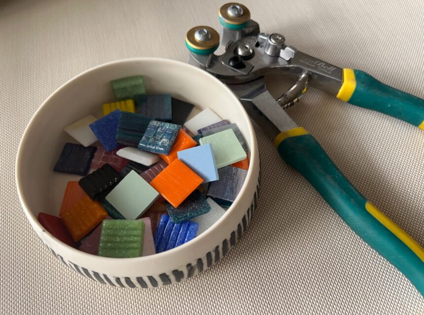 A bowl of colourful mosaic tiles sits beside a green-handled tile nipper tool on a light-coloured surface.