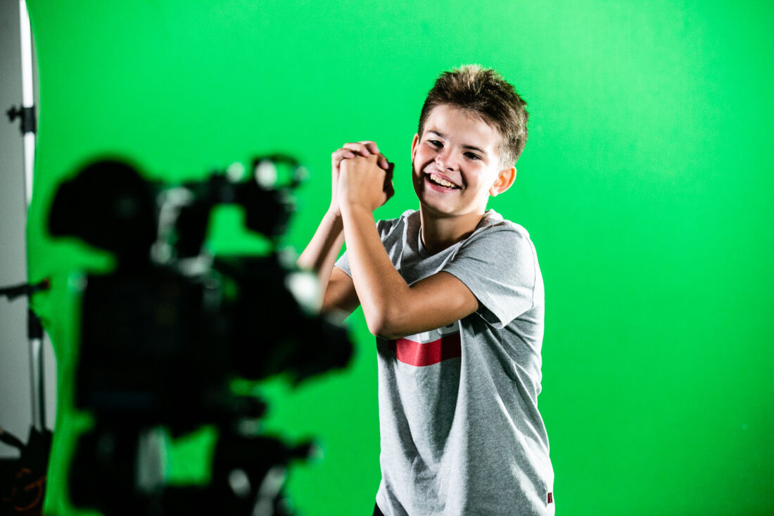 Smiling boy in a grey T-shirt stands in front of a green screen, clasping his hands together, with a camera on a tripod visible in the foreground.