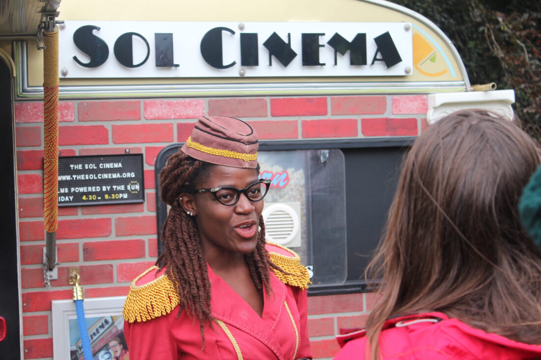 A woman in a red uniform with gold trim and glasses stands outside a caravan labelled Sol Cinema, speaking to another person whose back is to the camera.