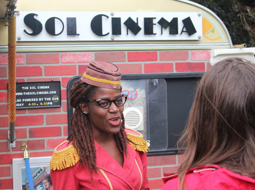 A woman in a red uniform with gold trim and glasses stands outside a caravan labelled Sol Cinema, speaking to another person whose back is to the camera.