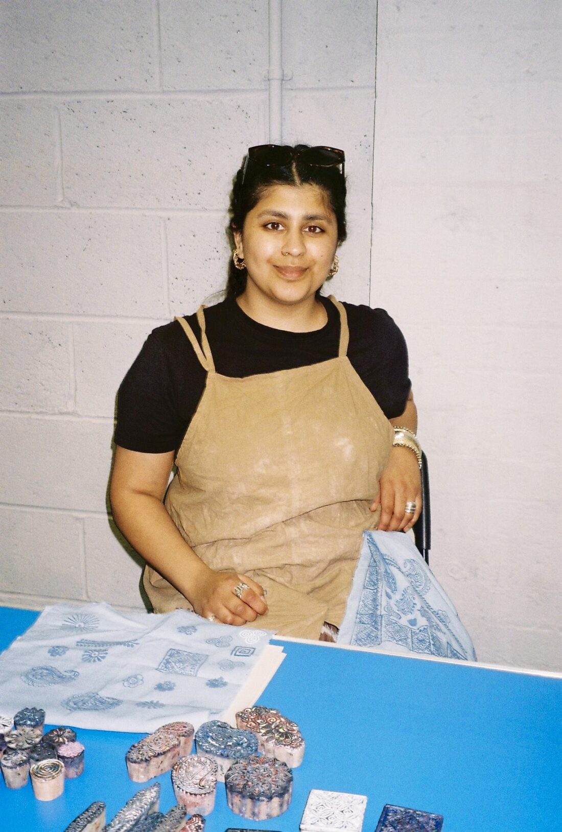 A person wearing an apron sits at a blue table displaying carved stamps and printed fabric patterns, smiling at the camera.