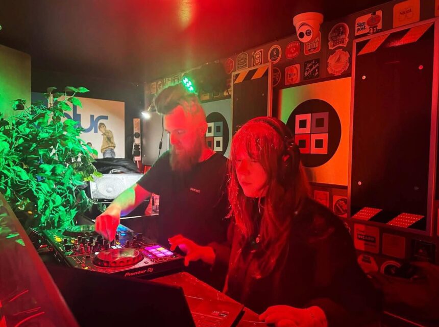 Two DJs perform together in a dimly lit booth with colourful lights, headphones on, surrounded by stickers and equipment. A leafy plant is visible to the left.