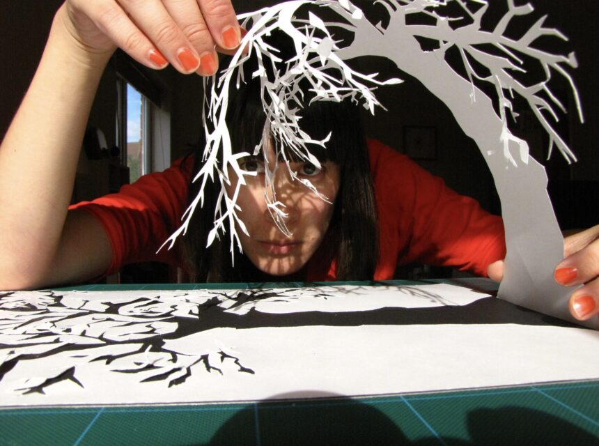 A person with orange nail varnish holds a delicate paper cut-out of a tree, carefully positioning it above a table with its shadow and silhouette also visible.