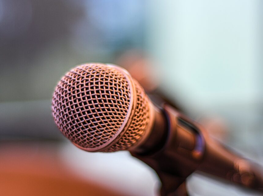 Close-up of a silver microphone with a mesh head and black handle, positioned at an angle against a blurred background with soft blue and brown tones.