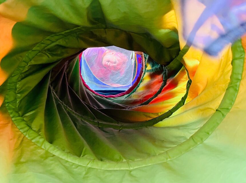 Brightly coloured tunnel with spiralling green, yellow, and orange fabric, viewed from inside towards the circular opening, creating a vibrant abstract pattern and a glimpse of more colours at the far end.