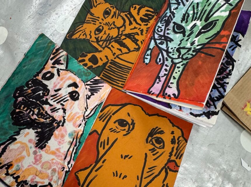 Four colourful artworks of cats and dogs drawn in bold black lines on orange, green, red, and teal paper are displayed on a silver spotty surface, showing the animals in playful and expressive poses.