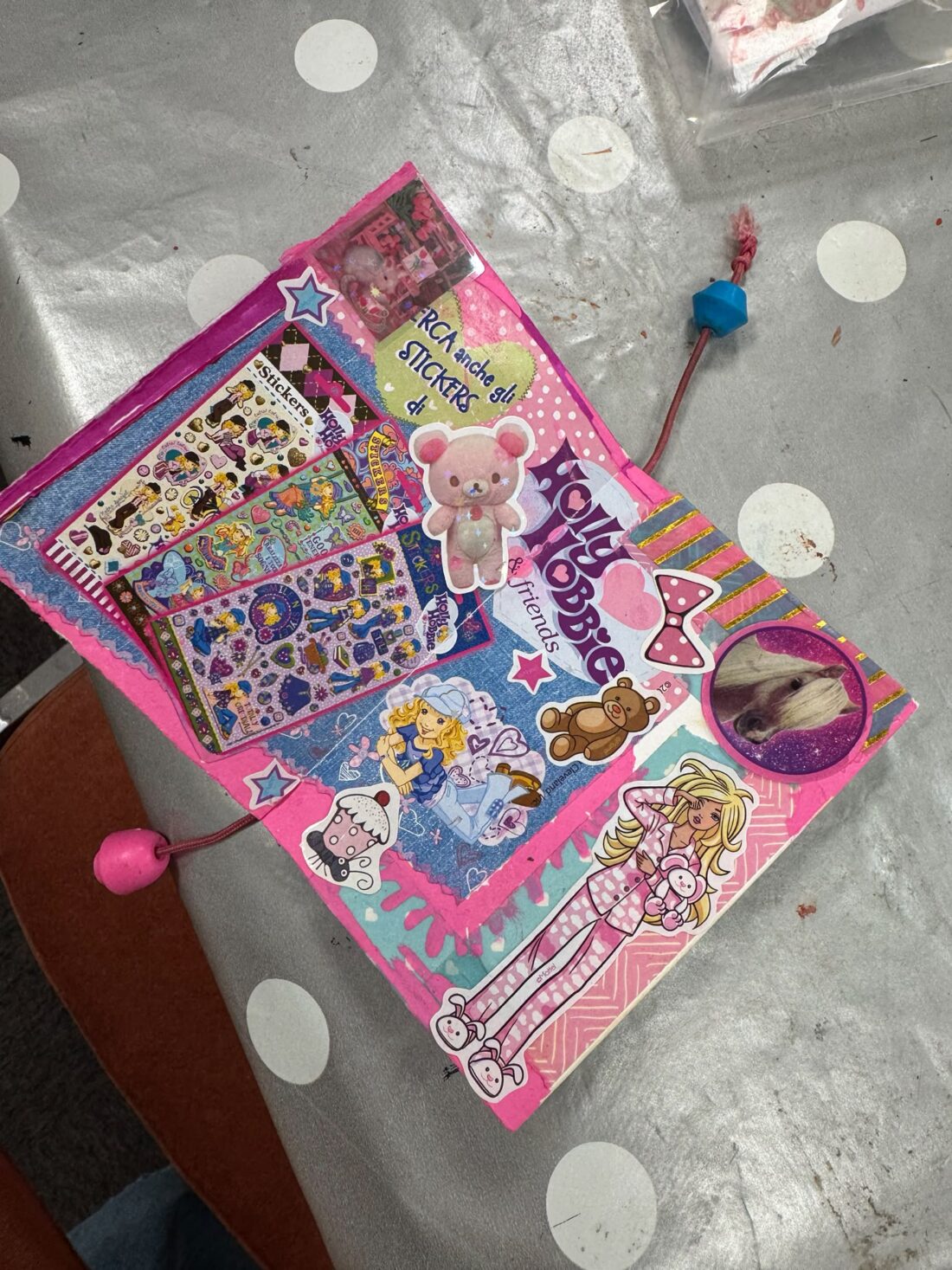A pink scrapbook page decorated with colourful stickers, a photo of a guinea pig, Barbie drawings, and cartoon images, lying on a grey polka-dotted tablecloth. Two tassels with beads are attached to the page.