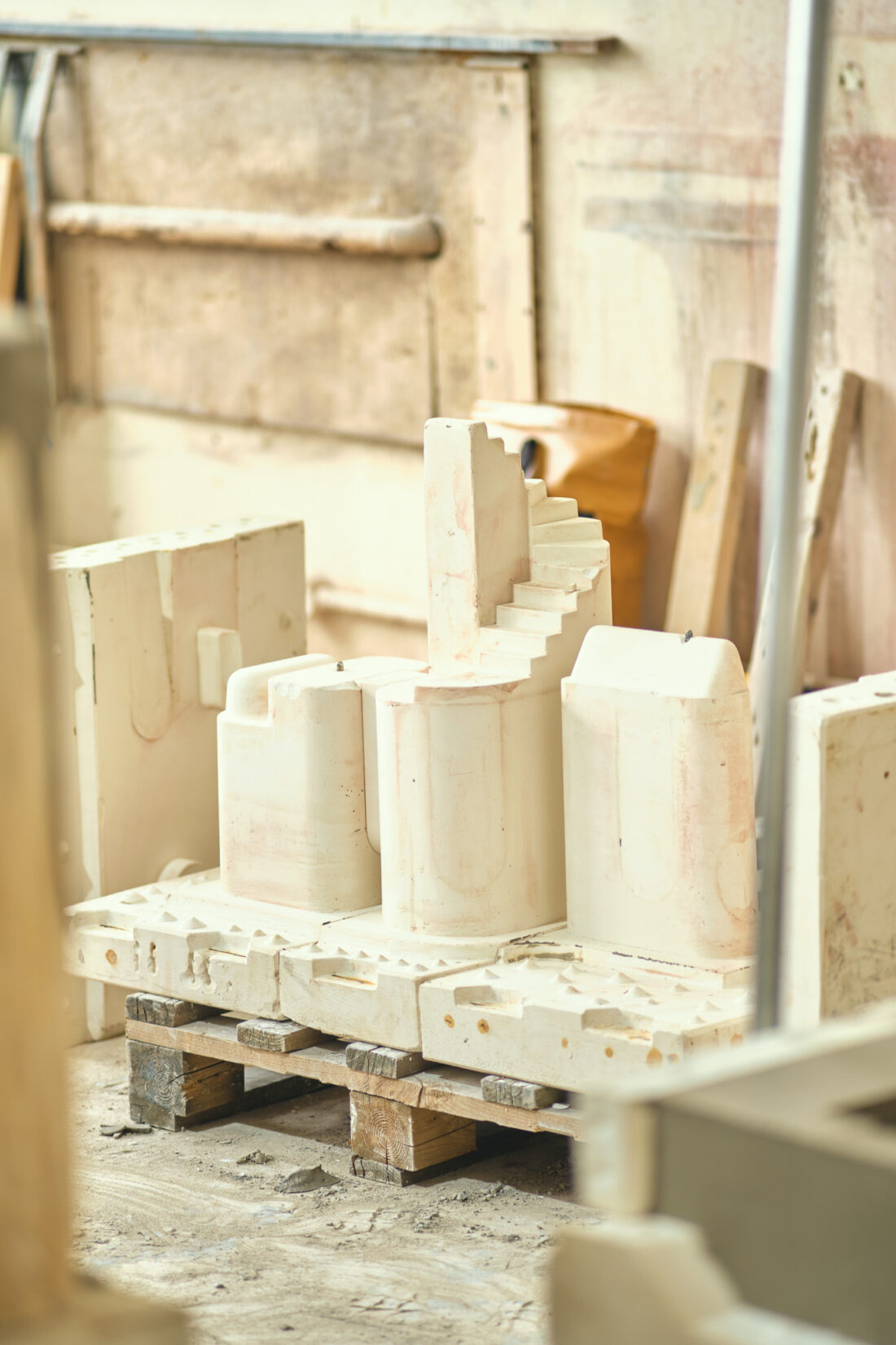 Sculpted plaster architectural models, including one with stairs, are displayed on wooden pallets in a workshop with various tools and materials in the background.