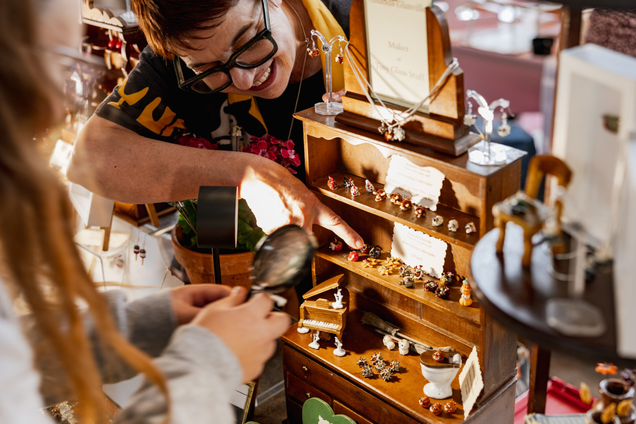 A person examines small jewellery pieces with a magnifying glass at a wooden display, while the seller smiles and points out items. The display holds various colourful rings and miniatures.