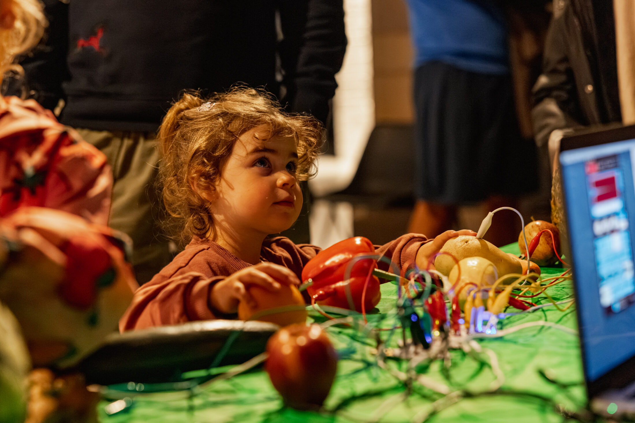 A young child sits at a table with fruit and vegetables connected by wires, looking up with curiosity. A computer screen is visible nearby, and other people are in the background.