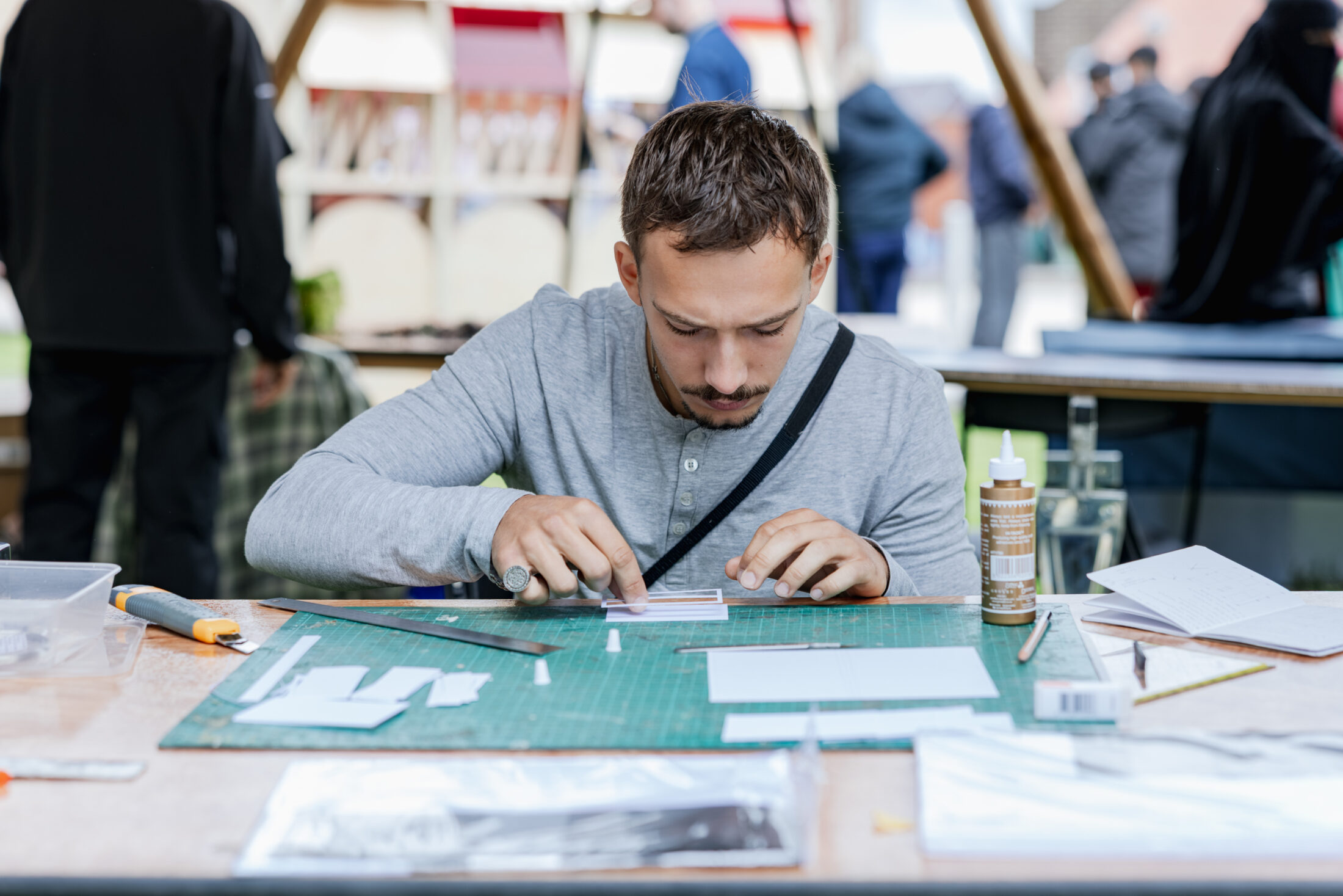 A man sits at a table, focusing intently as he works on a craft project with paper, a cutting mat, and tools. Other people and stands are visible in the blurred background.