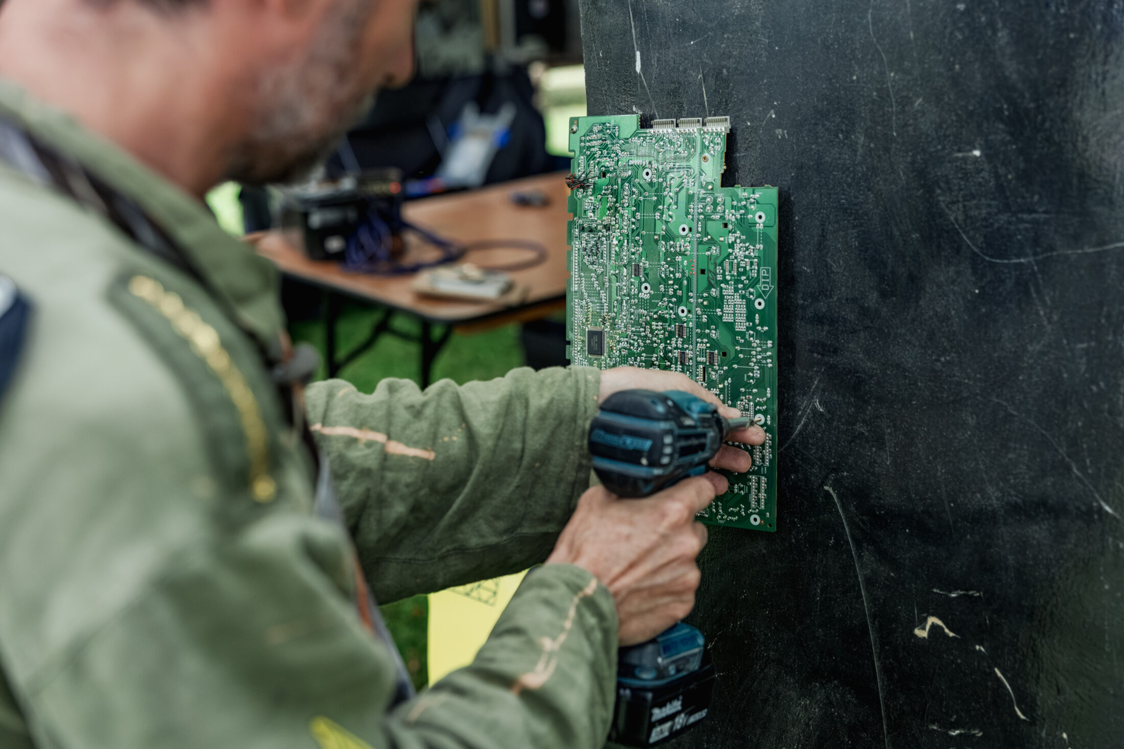 A person in a green jacket uses a power drill to attach a green circuit board to a dark vertical surface. A table with electronics is seen in the blurred background.