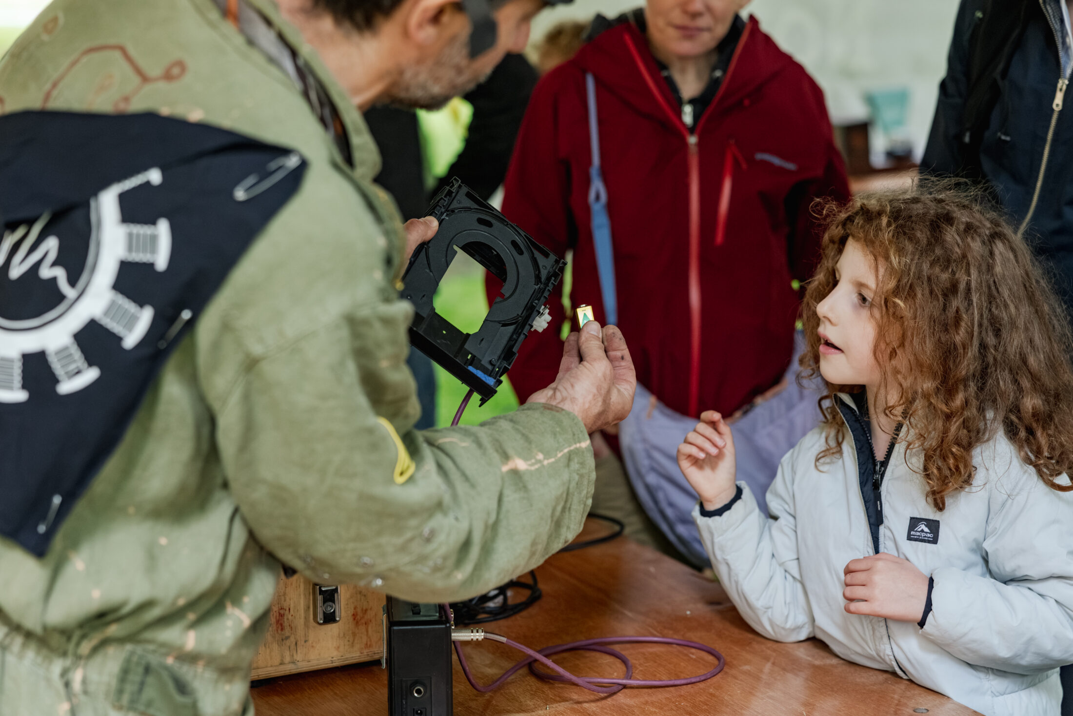 An adult shows electronic parts to a curious young girl at a table, whilst other people watch in the background. The setting appears interactive and educational.