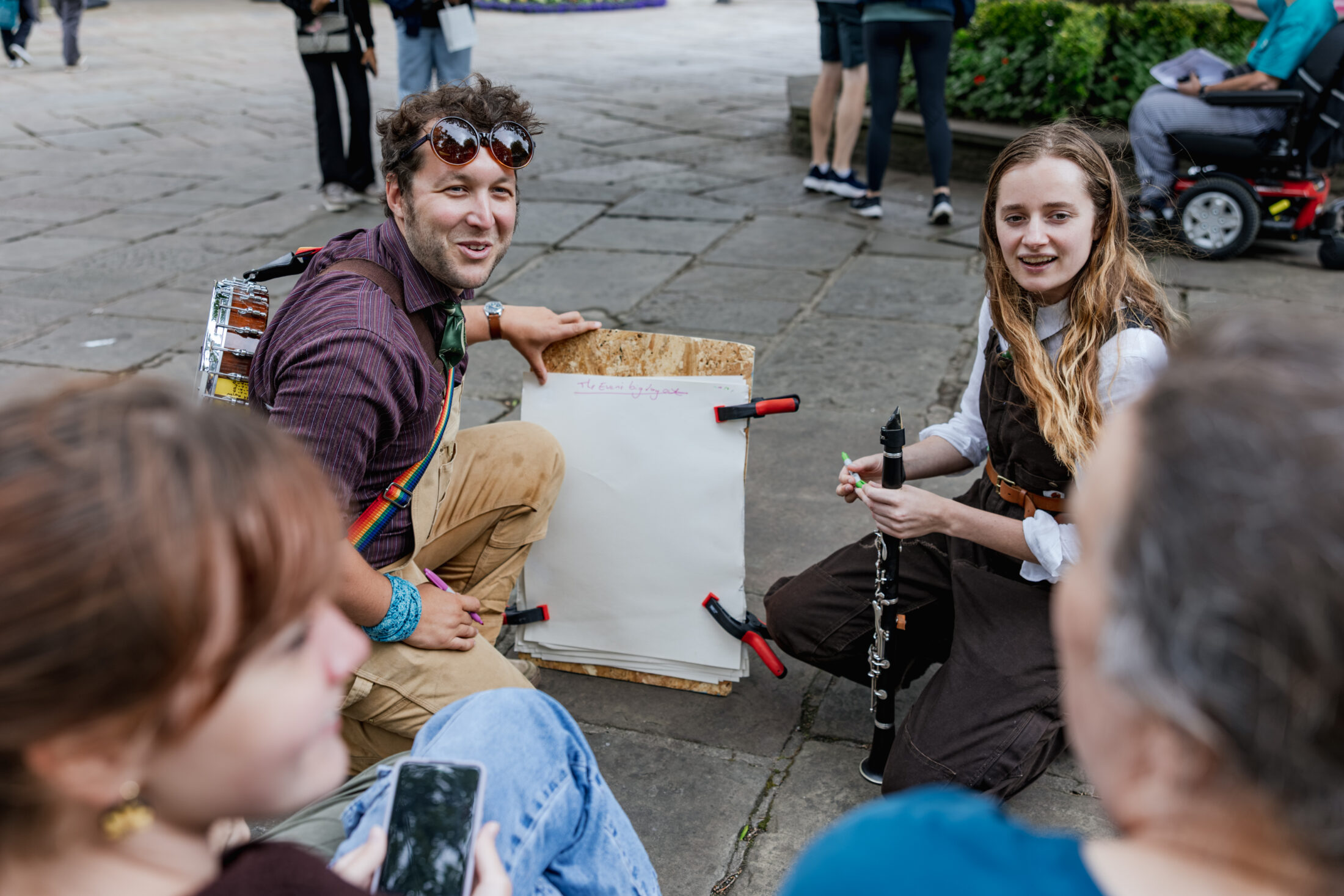 Two people kneel on the pavement with a blank pad and musical instrument, engaging with two seated individuals in an outdoor setting. A person in a wheelchair and others are visible in the background.