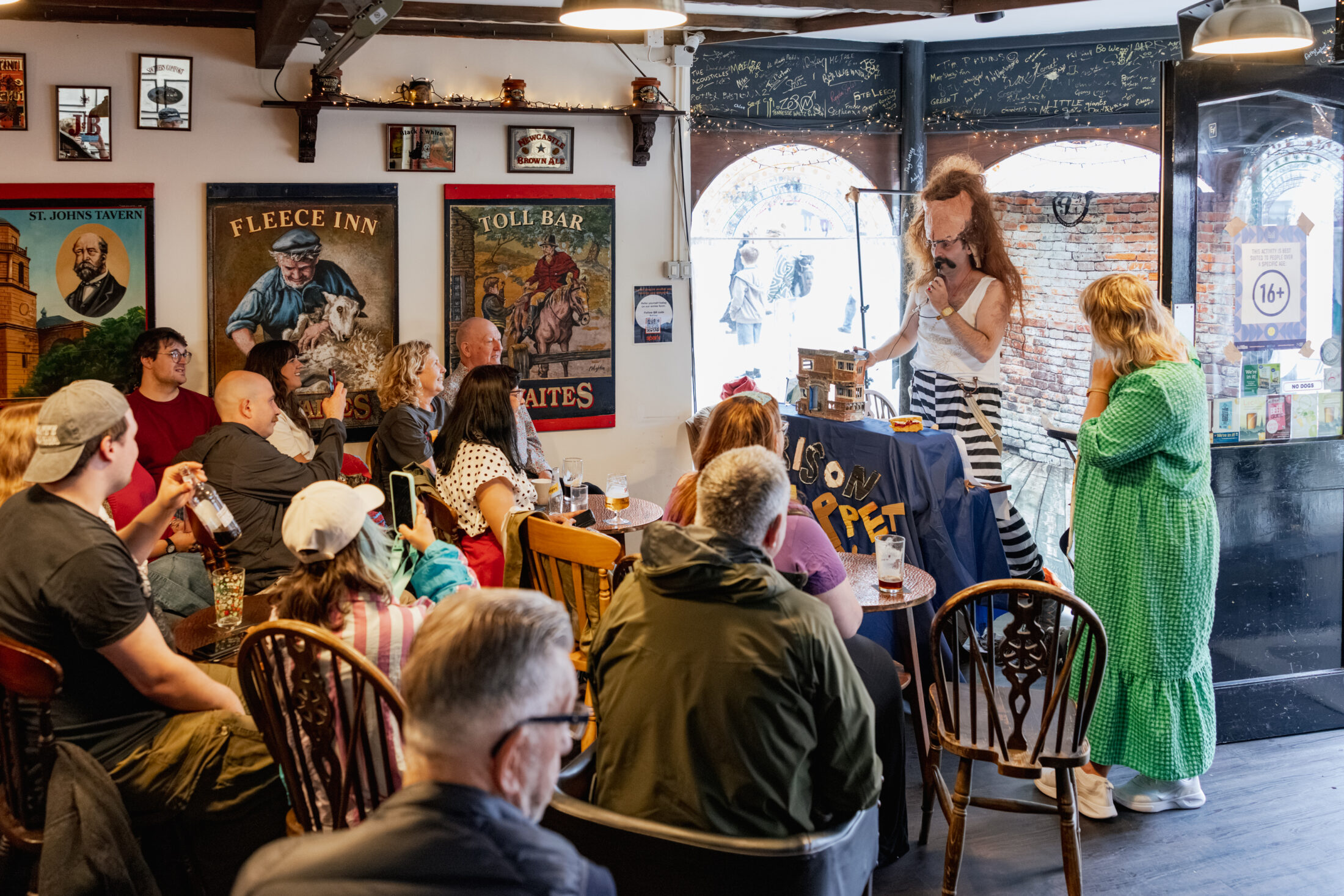 A performer in costume entertains a seated audience at a cosy pub. Posters line the walls, drinks are on tables, and sunlight streams in through arched windows and an open door. A sign on the door reads 16+.