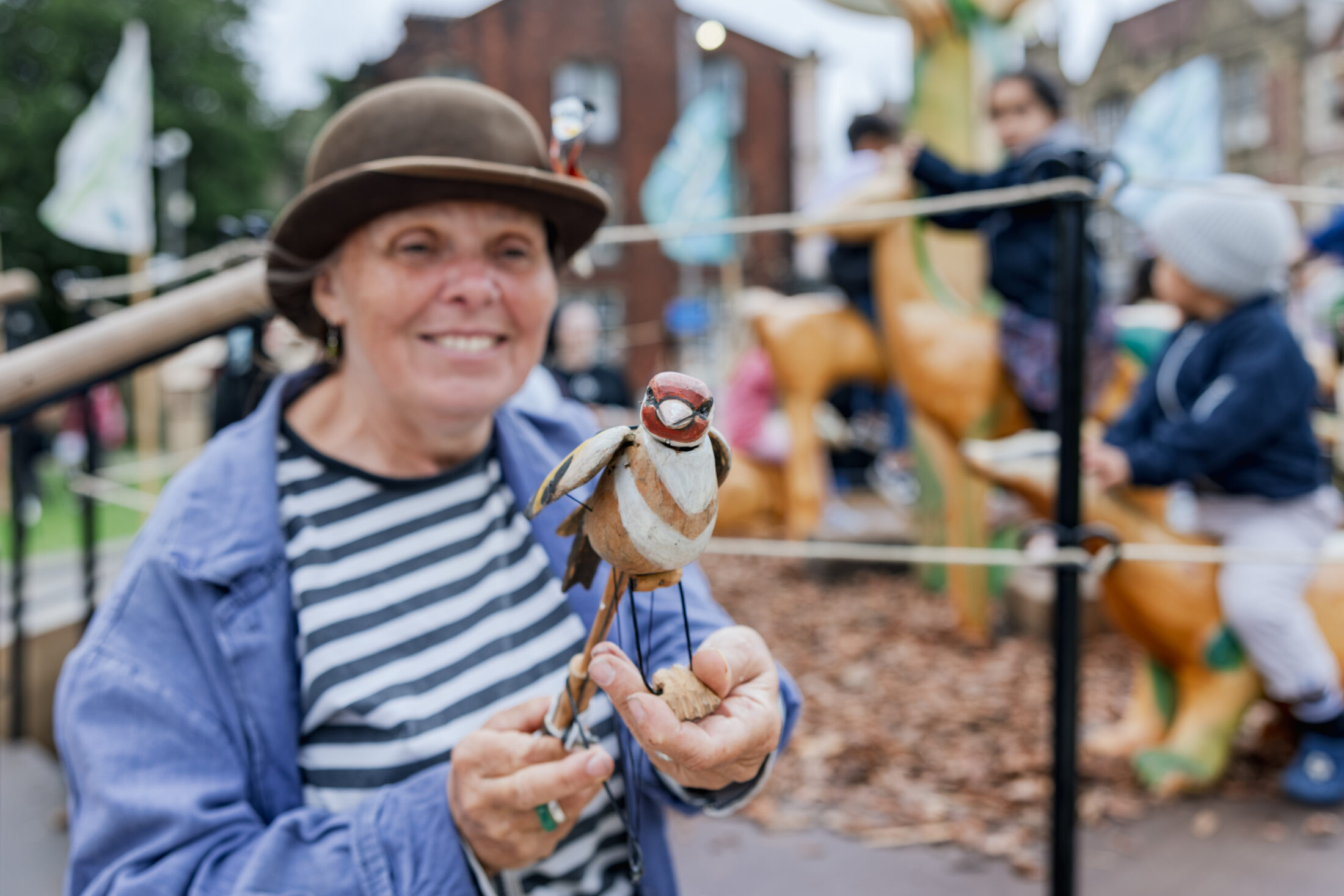 A smiling woman in a hat and striped shirt holds a small bird puppet outdoors at an event. Children and animal sculptures are visible in the blurred background.