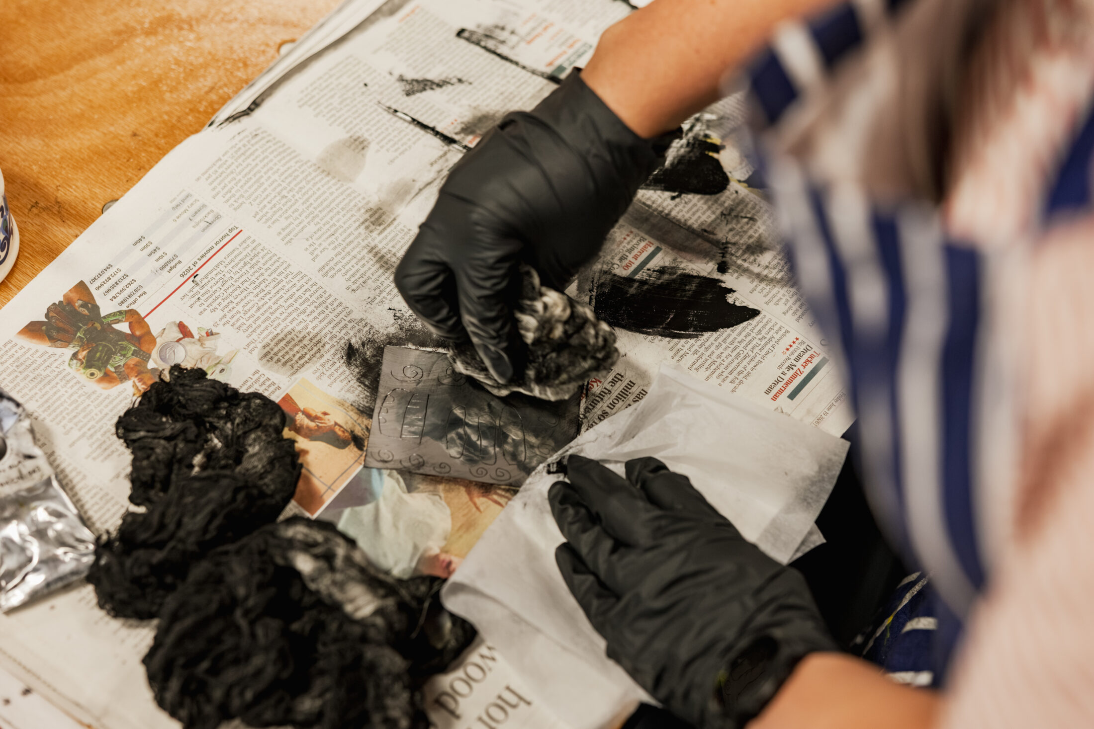 A person wearing black gloves cleans a small, dark photo plate on a newspaper-covered table, surrounded by used paper towels and smudges of black ink or paint.