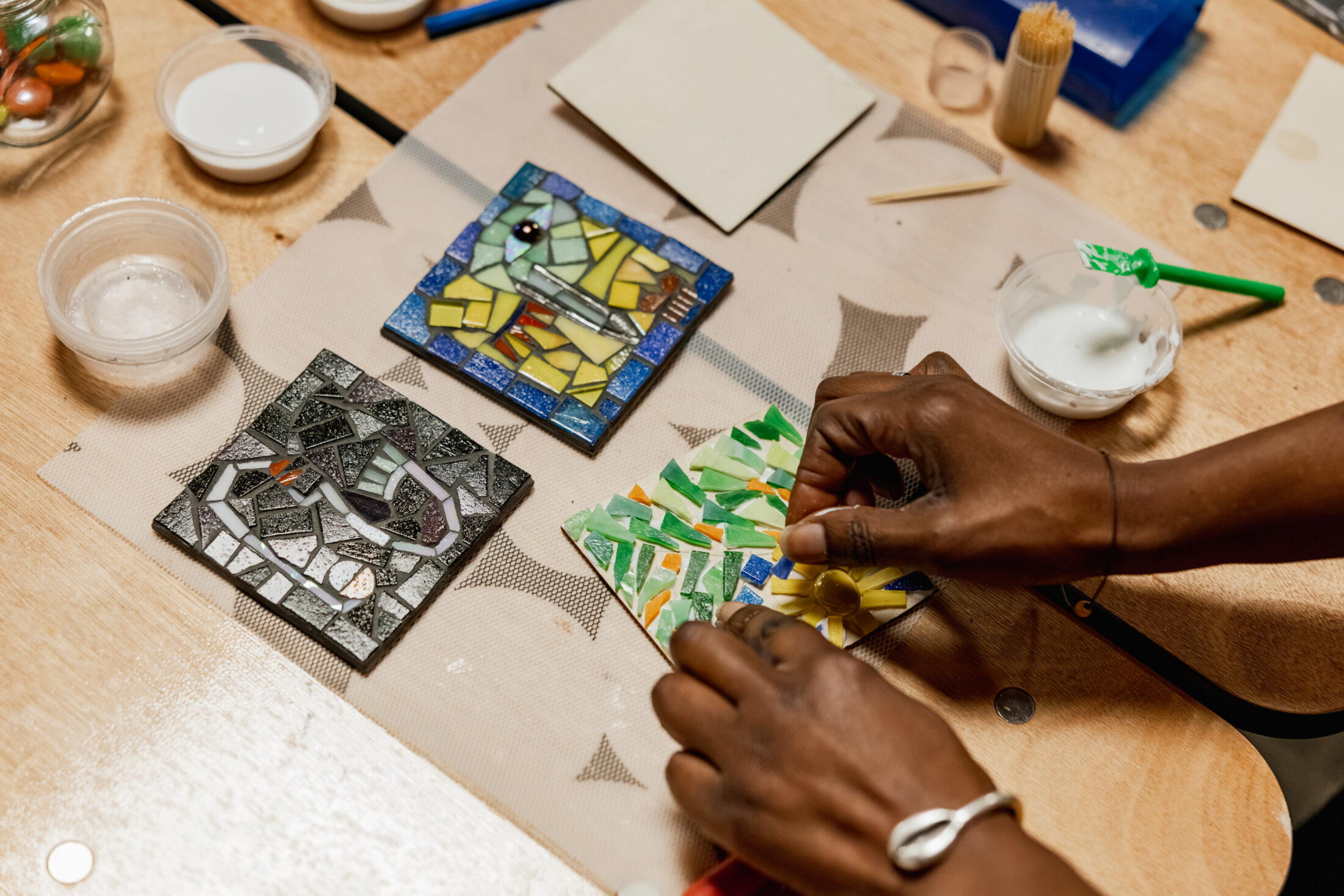 A person arranges colourful mosaic tiles on a square base, with two finished mosaic artworks nearby on a wooden table, surrounded by craft supplies.