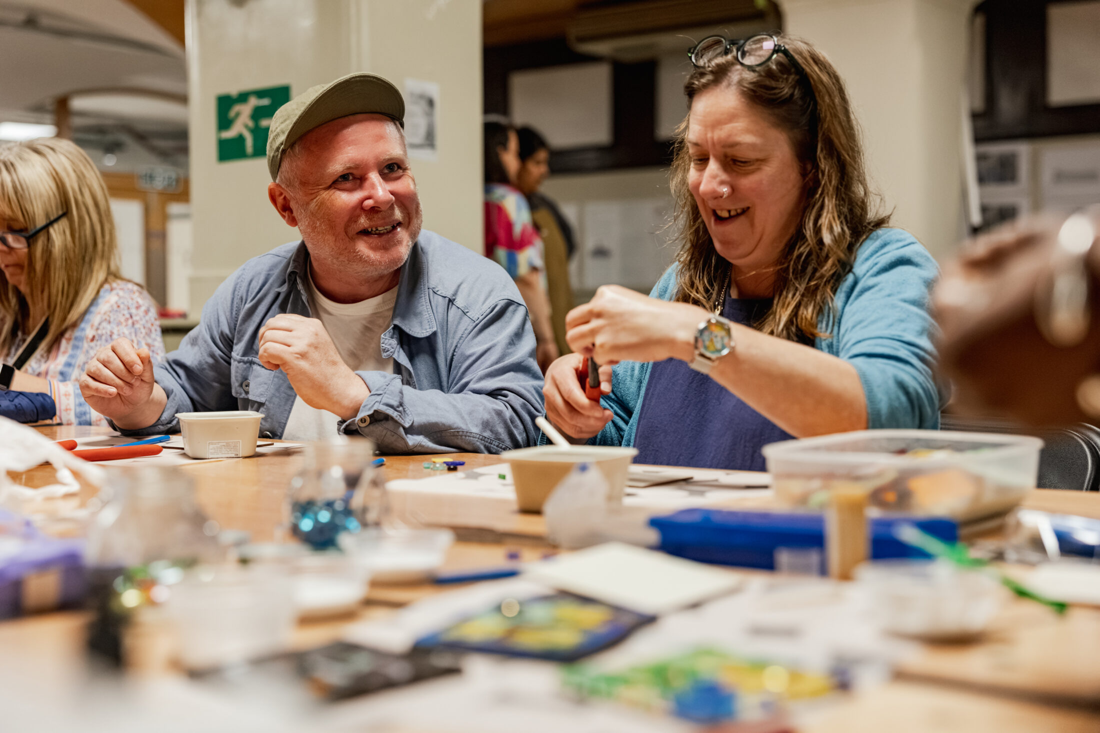 Two people sit at a table covered with craft supplies, smiling and working on art projects together in a lively, indoor setting. Others are also engaged in activities in the background.