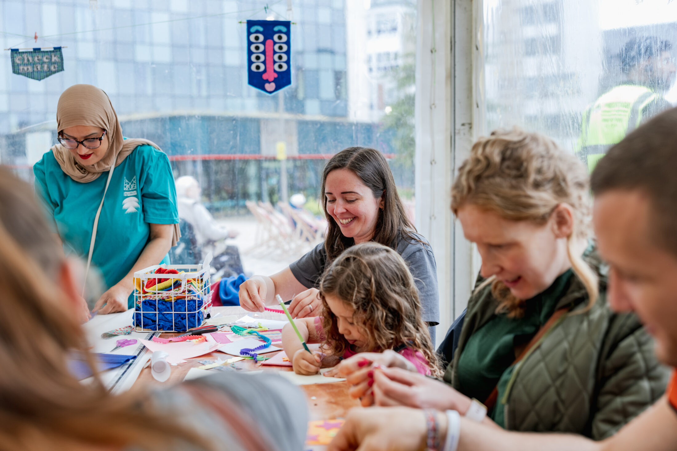 Adults and children sit around a table, happily crafting with colourful paper and art supplies in a bright room with large windows.
