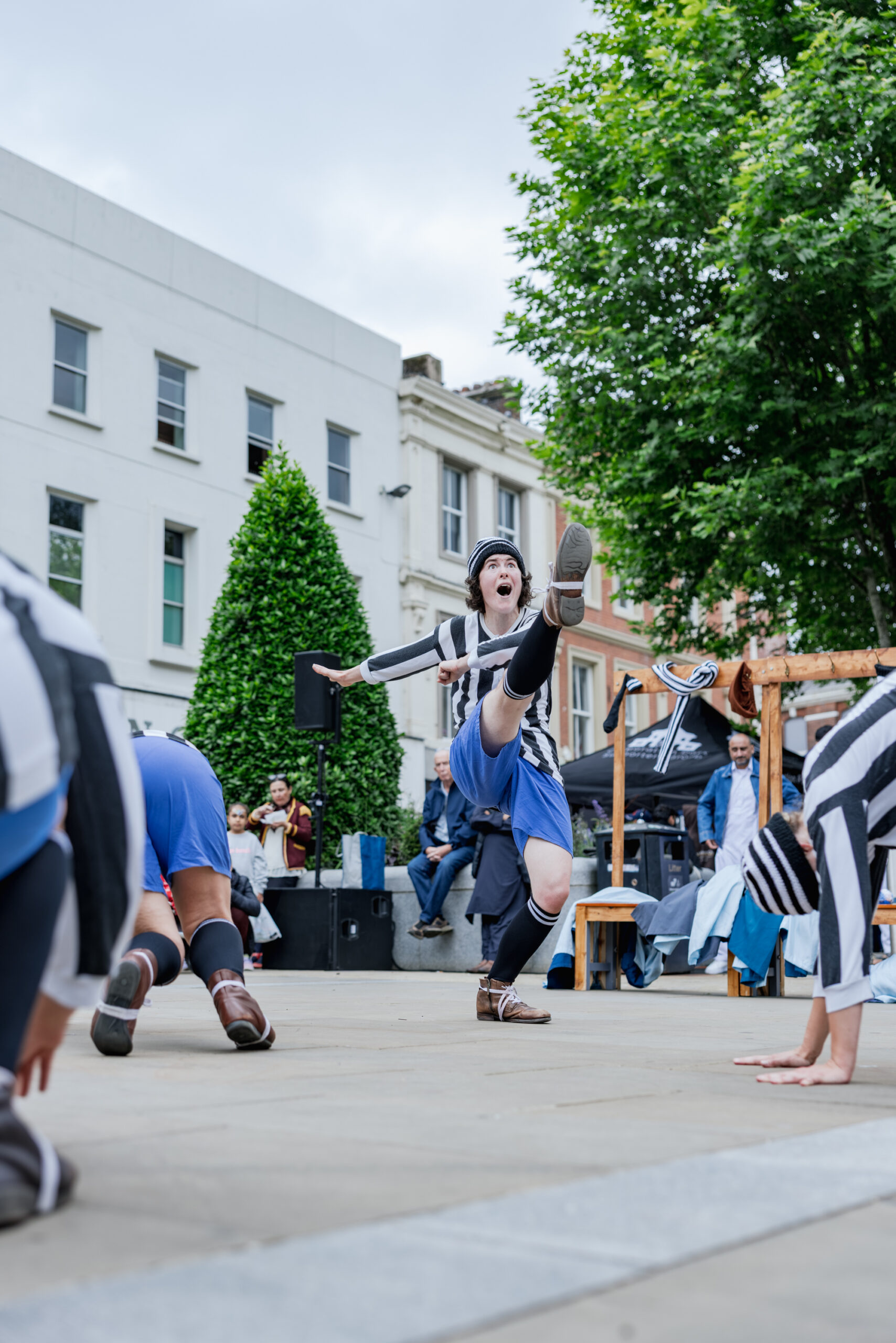 A street performer in a blue skirt, striped shirt, and cap energetically kicks one leg high during an outdoor show, as others perform nearby. Spectators watch from benches.