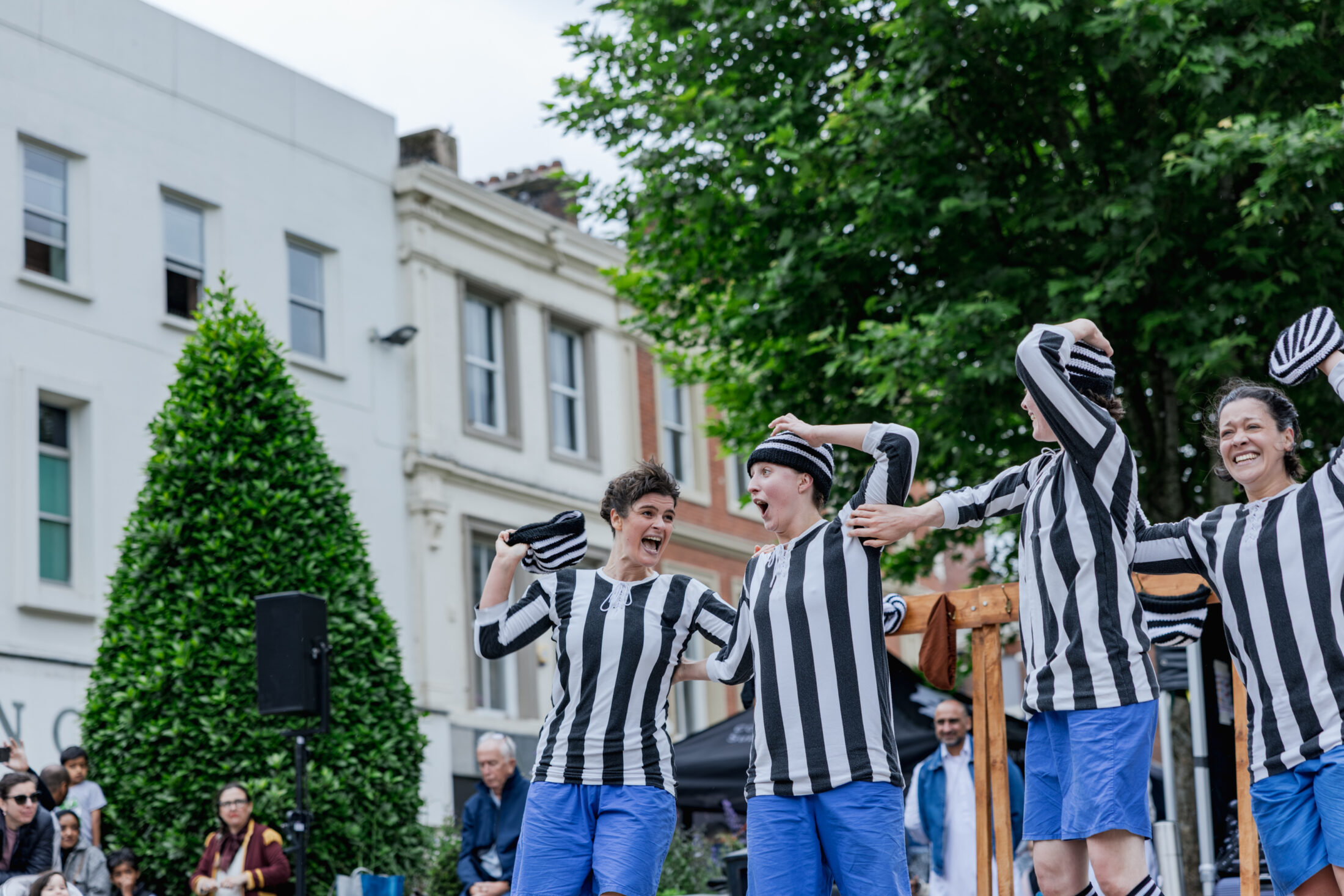 Four performers in black-and-white striped shirts and blue shorts dance and smile on a street, with a crowd watching in the background and trees nearby.