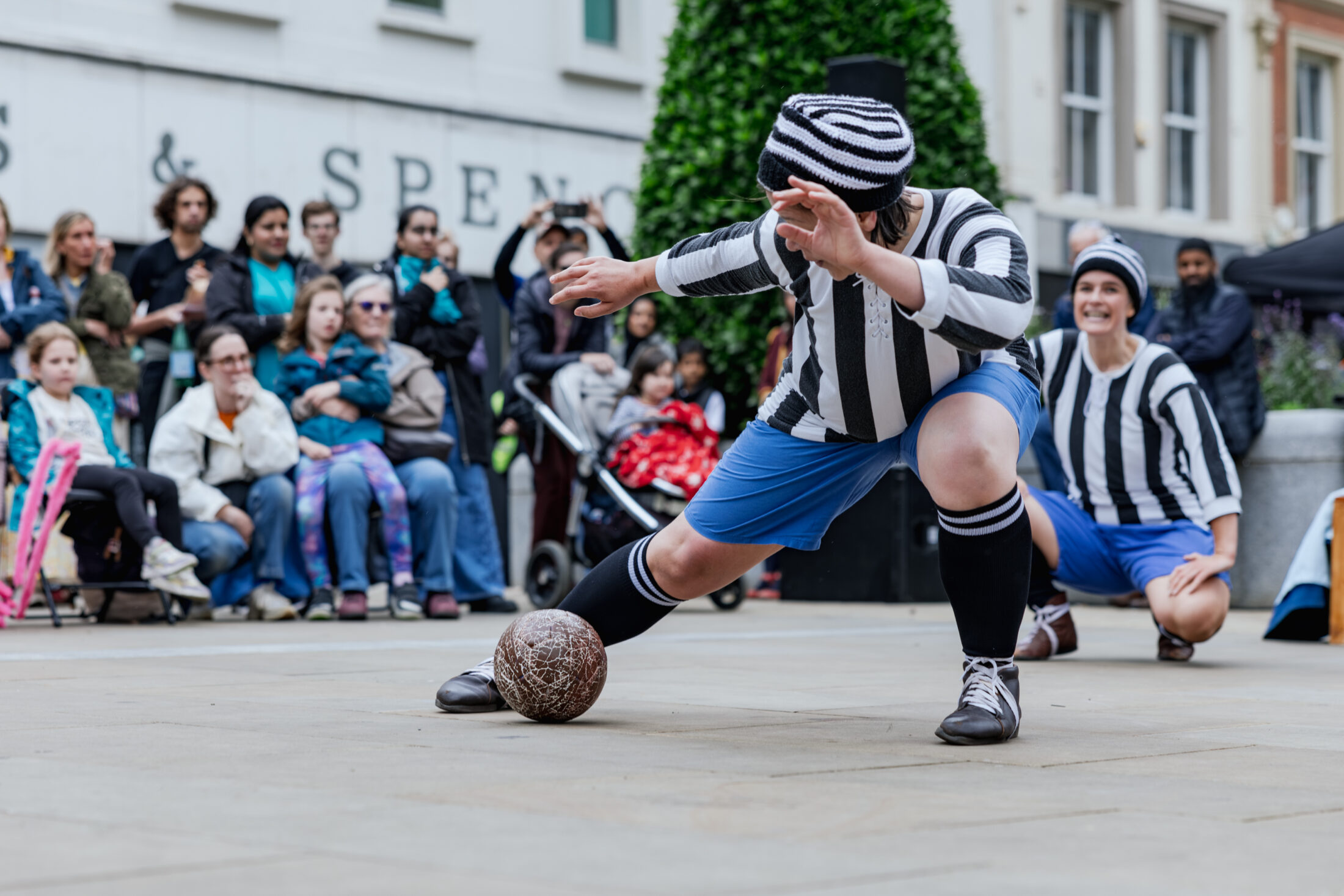 A performer in a black-and-white striped outfit and blue shorts poses dramatically with a vintage football, while a crowd watches the street performance. Another performer in similar attire smiles in the background.