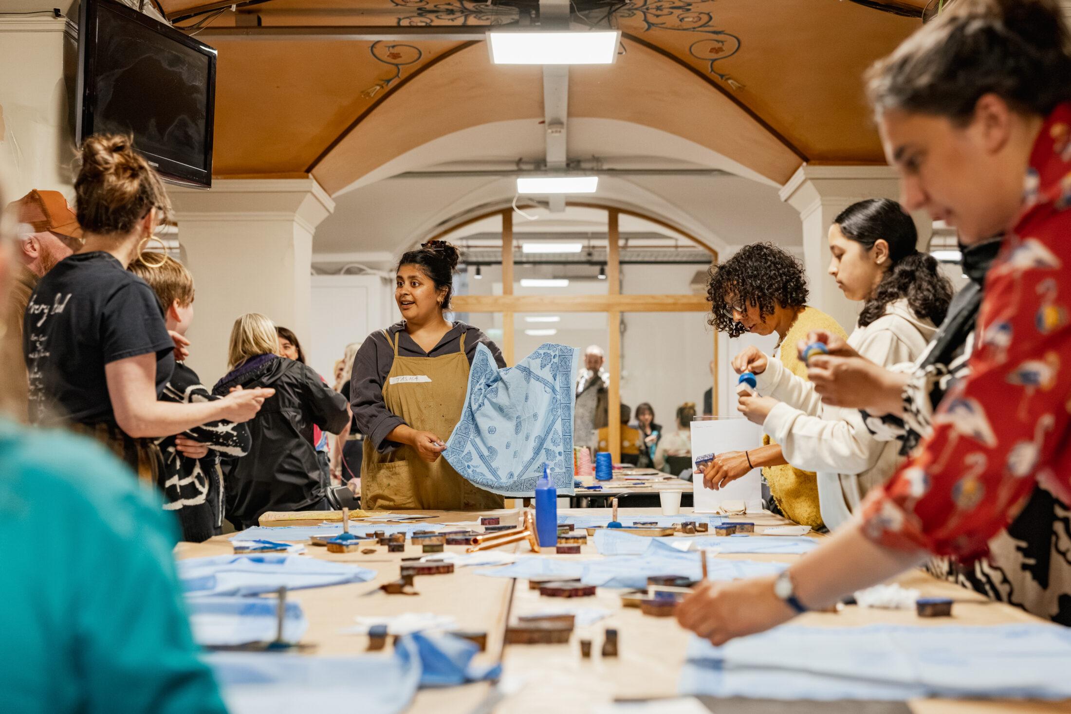 A group of people participate in an indoor arts and crafts workshop, working together at long tables covered with blue fabric and supplies.