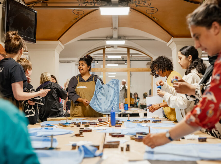 A group of people participate in an indoor arts and crafts workshop, working together at long tables covered with blue fabric and supplies.