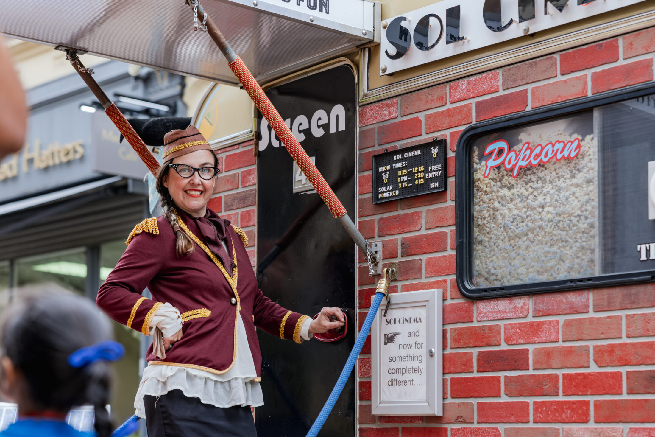A smiling woman in a vintage-style uniform stands by a popcorn machine on a brick wall. She wears glasses, a hat, and a maroon jacket with gold trim. A board with film times and a sign are visible nearby.