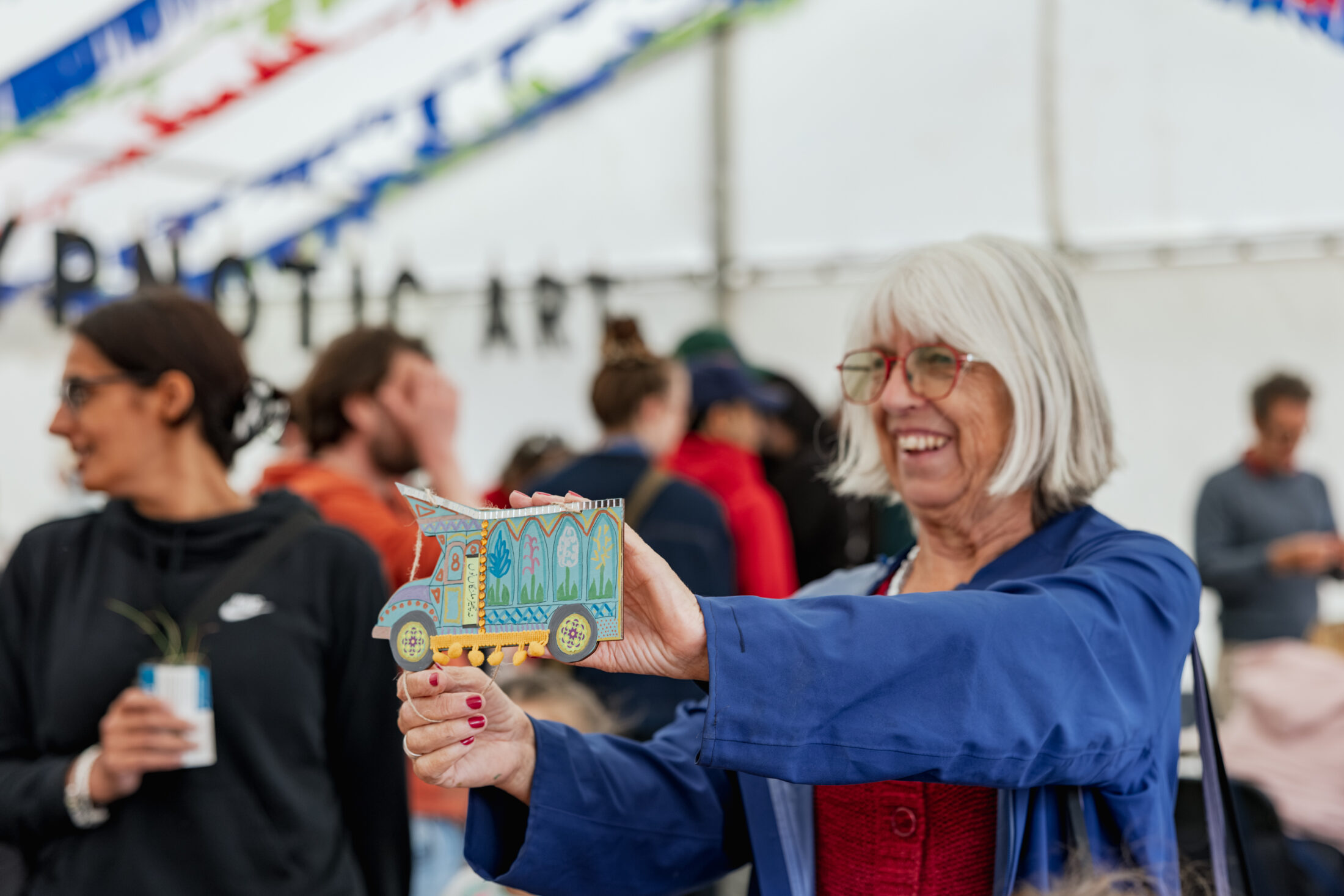 An older woman with grey hair and glasses smiles whilst holding a colourful cardboard cut-out of a lorry at a lively indoor event with people and festive bunting in the background.