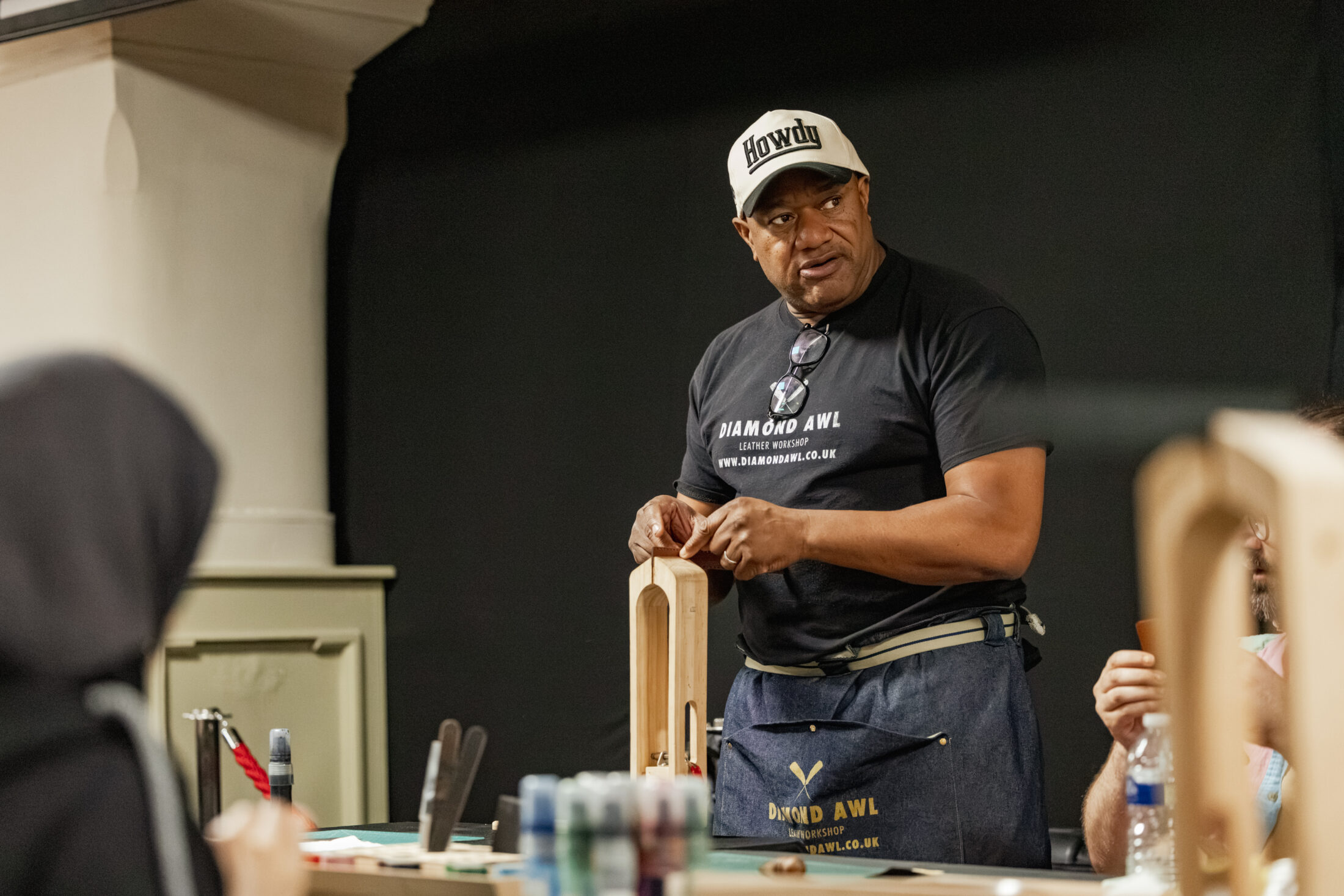A man wearing a Howdy hat and apron stands at a workbench, appearing to teach or demonstrate a craft project to others in a workshop setting.