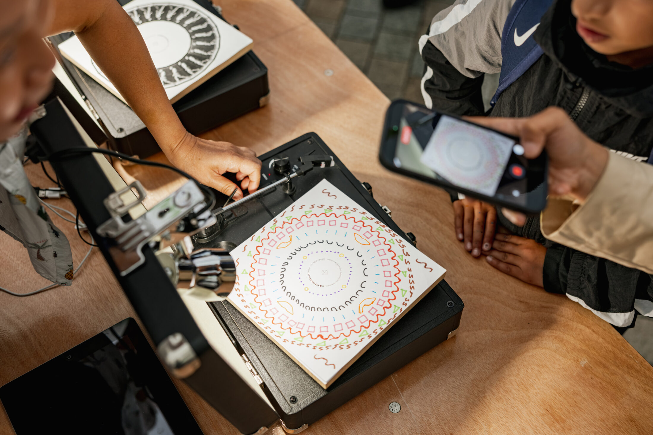 Two people use a drawing machine to create circular geometric art on paper, while one person photographs the process with a mobile phone. A child watches nearby.