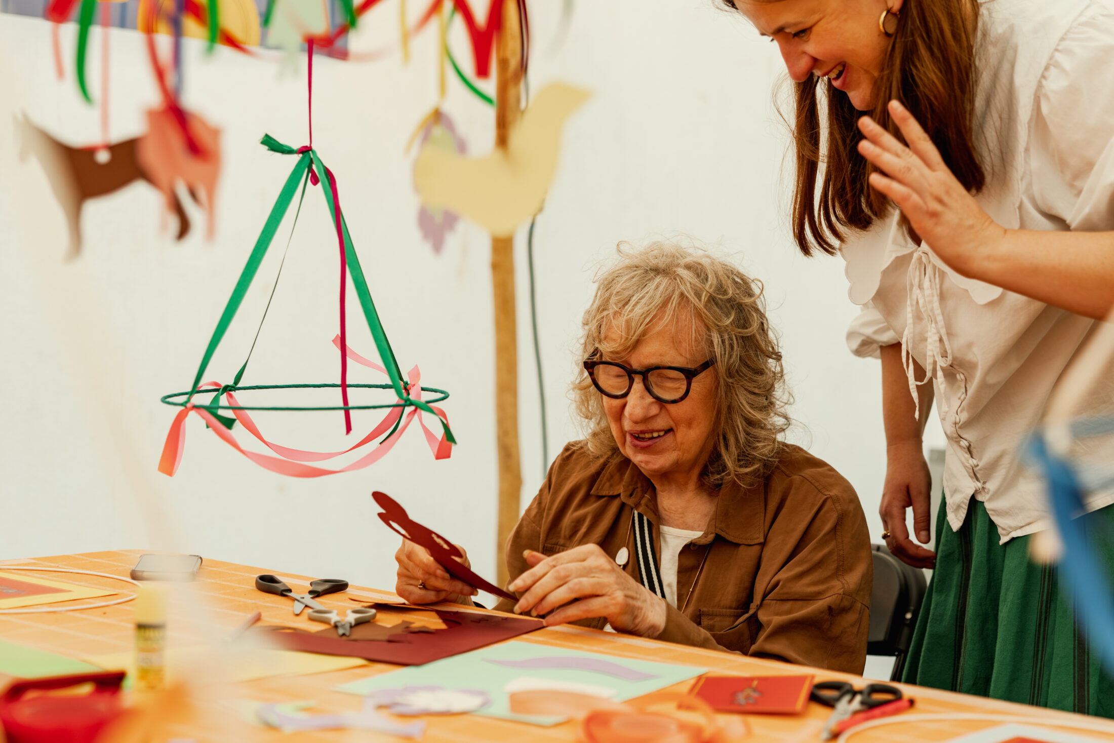 An older woman with grey hair and glasses smiles whilst doing crafts with paper animals, as a younger woman stands beside her, also smiling, at a decorated table.