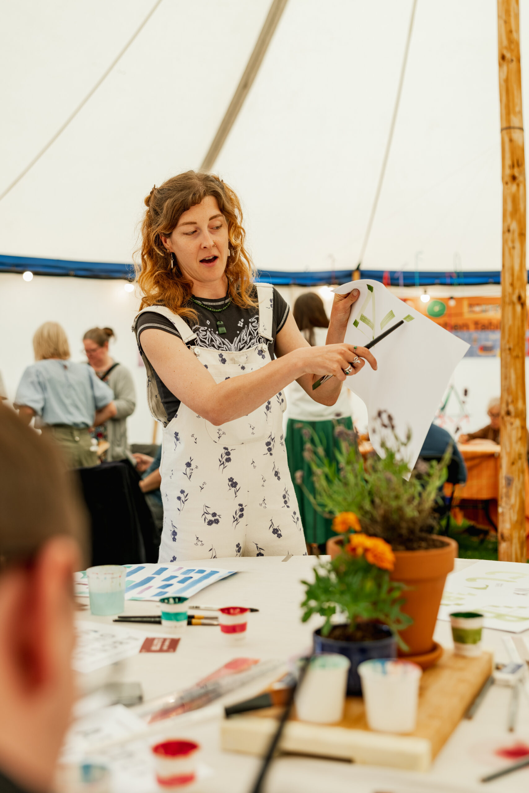 A woman stands in a marquee, holding up and explaining a sheet of paper with painted designs to people seated at a table with art supplies and potted flowers. Other attendees are visible in the background.