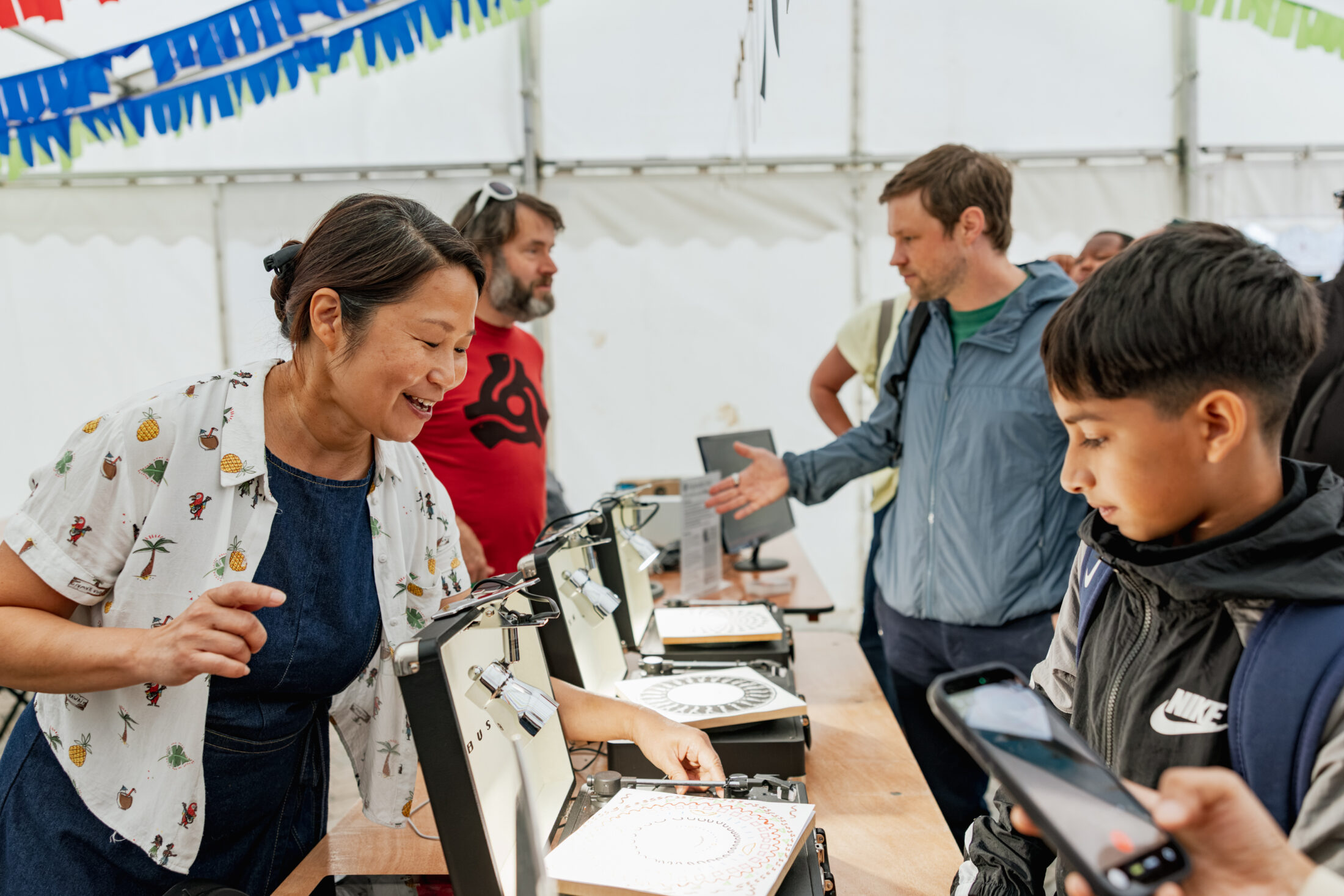 A woman demonstrates a drawing machine to a boy at a crowded stand, while other people observe and interact with similar machines in a brightly lit marquee.