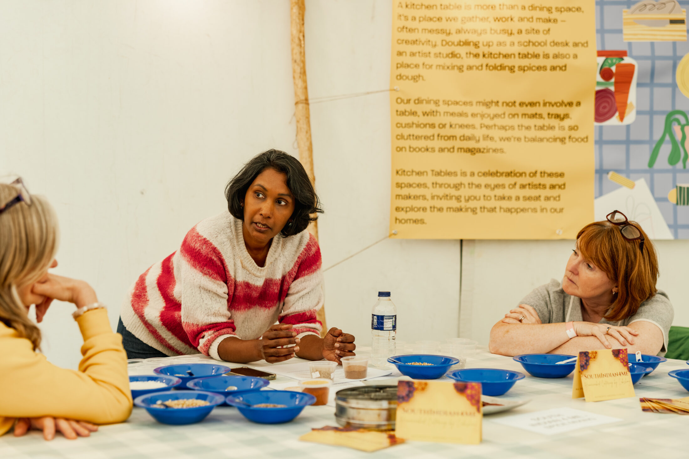 Three women sit at a table with blue bowls, engaged in discussion. One woman stands, gesturing as she speaks. A poster and books are visible in the background.