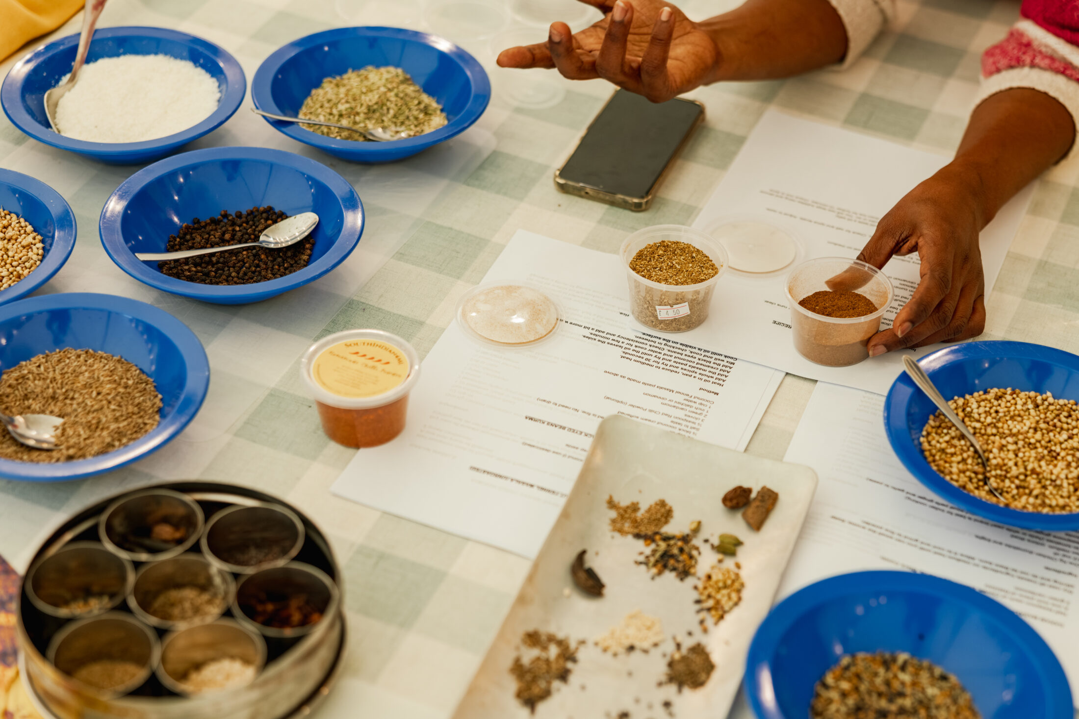 A person's hands arrange spices and grains in blue bowls and containers on a table covered with papers and a mobile, suggesting a cooking or recipe preparation scene.