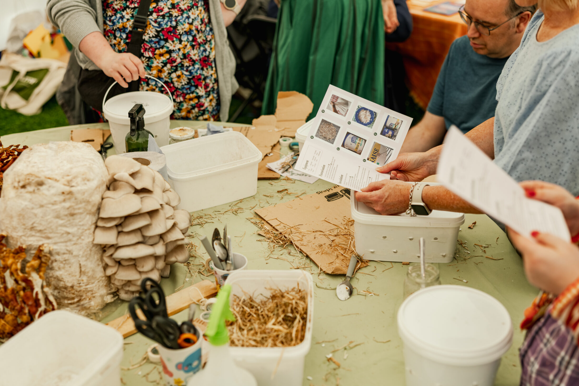 Several people gather around a table covered with mushrooms, straw, scissors, and containers, following printed instructions, likely participating in a hands-on mushroom growing workshop.