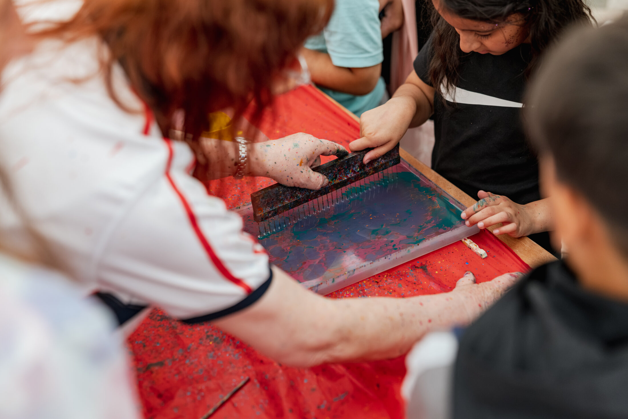 A child and an adult use a squeegee to spread paint on a screen for screen printing at a red table, with other children nearby.