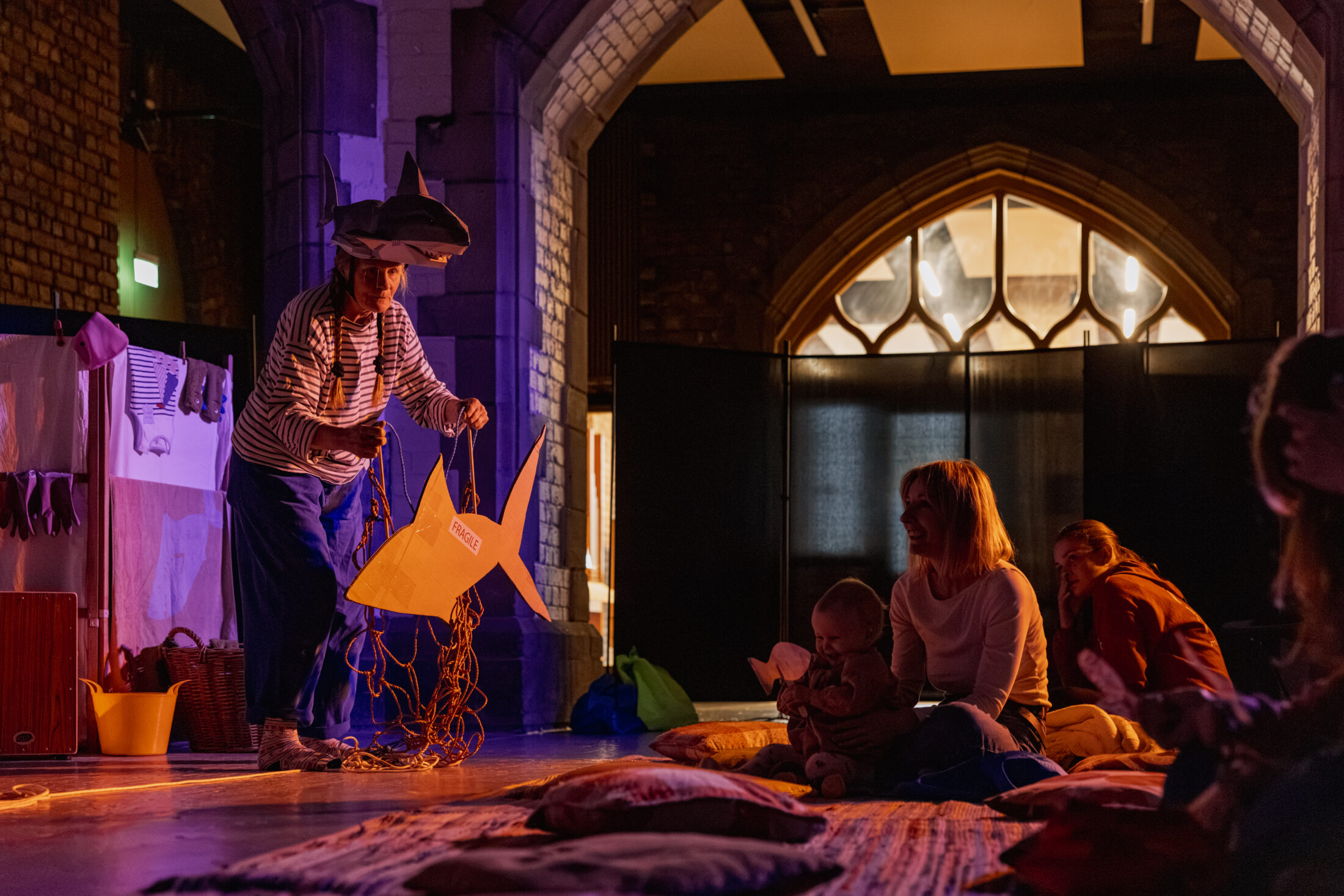 A performer in a striped shirt and shark hat entertains a small audience, including a seated woman holding a baby, in a dimly lit room with arched windows and scattered cushions on the floor.