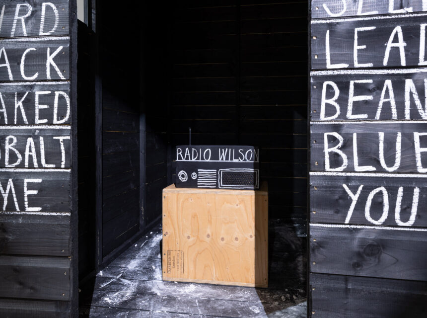 A small radio labelled RADIO WILSON sits on a wooden box in a dark room. Black walls on either side are covered with white chalk words like BIRD, JACK, BEANS, and BLUE.