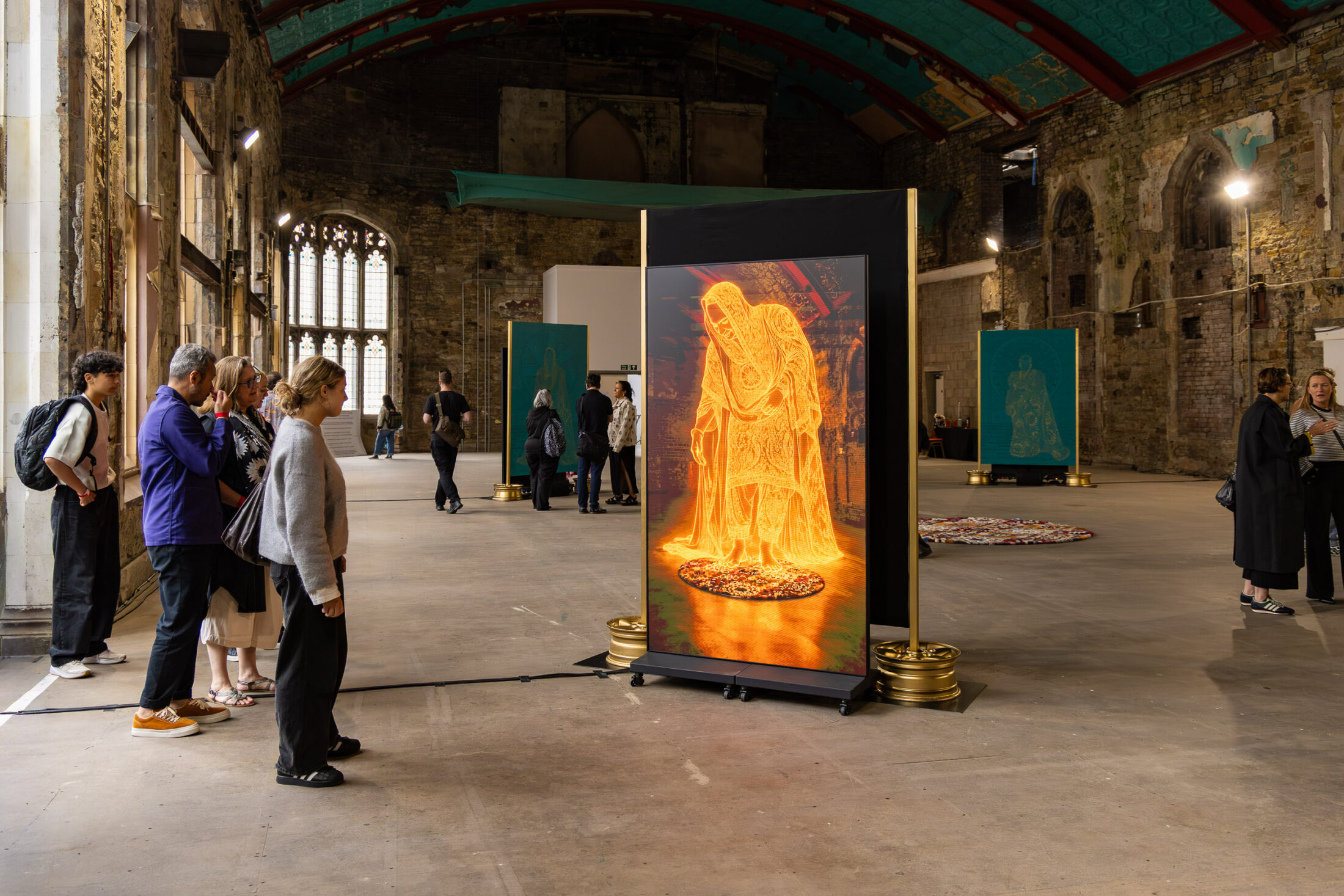 People view an illuminated digital artwork of a robed figure in a large historic hall with stone walls, arched windows, and other art pieces displayed throughout the space.