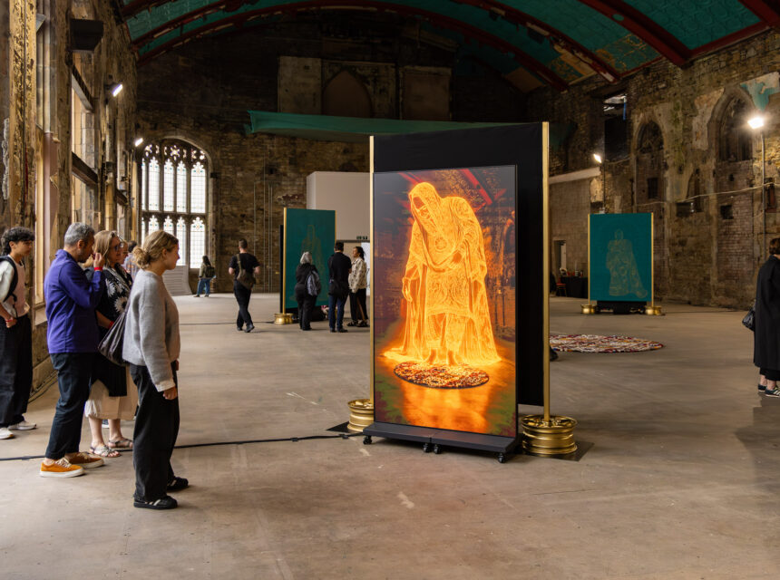 People view an illuminated digital artwork of a robed figure in a large historic hall with stone walls, arched windows, and other art pieces displayed throughout the space.